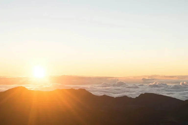 Sun rising over a mountain range with clouds below, casting a warm golden glow across the sky.