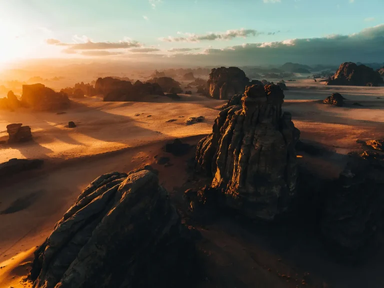 A vast rocky desert landscape at sunset, with large rock formations casting long shadows and a clear sky in the background.