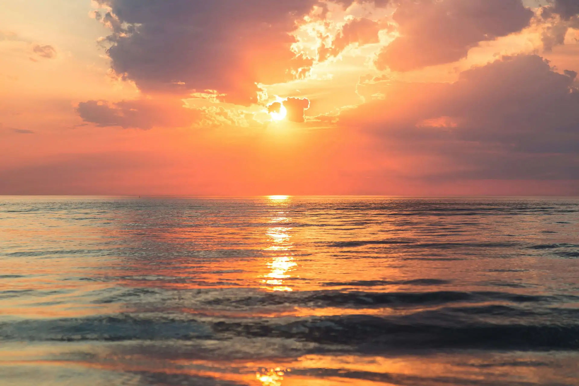 Sunset over a calm ocean, with orange and pink hues reflecting on the water and clouds partially covering the sun.